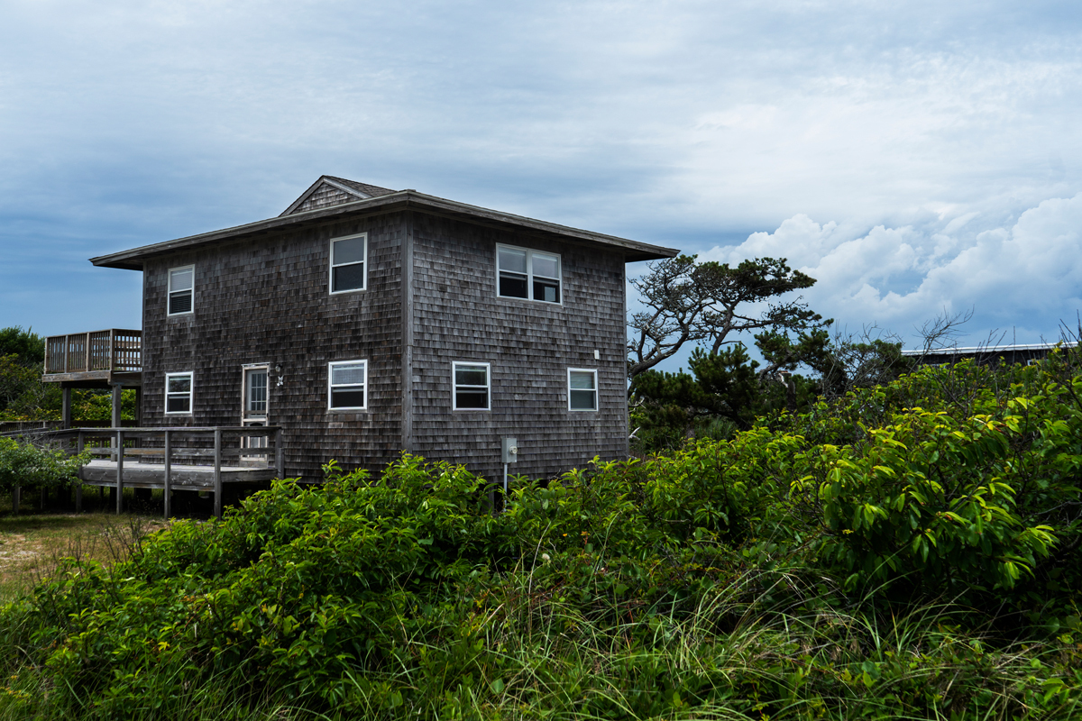 A two story beach house is visible, partially obscured by some lush vegetation. 