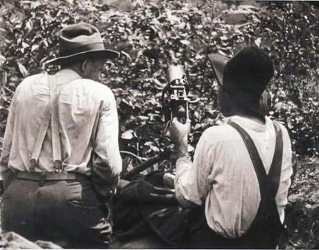 There are two men in white work shirts, suspenders and trousers facing away from the camera. They are kneeling behind a mound of earth. The man on the left has his head turned towards the man on the right, who mans a machine gun. Both are wearing wide-brimmed hats.