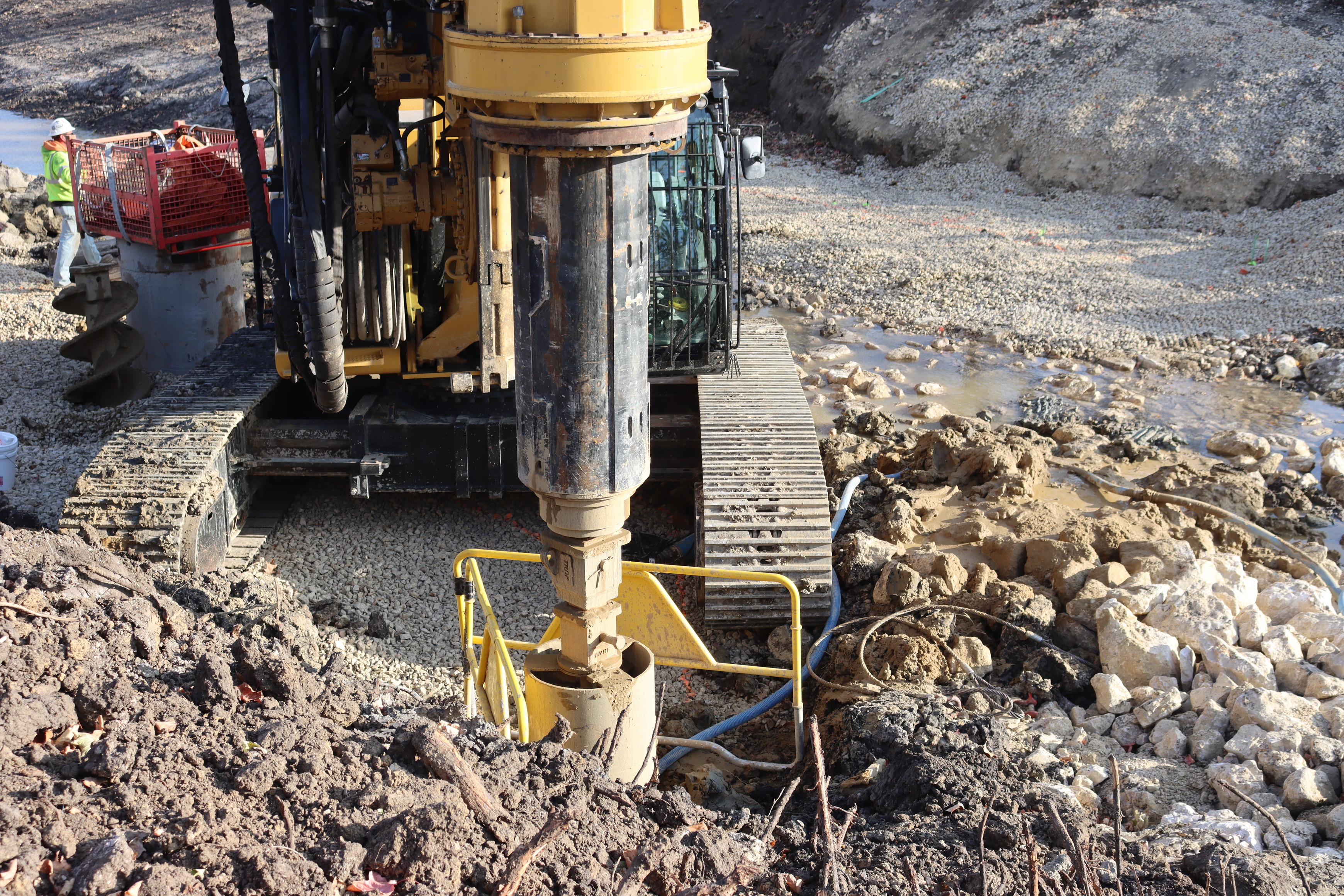 Large yellow construction equipment with drill attachment in center of image bores holes to be filled with concrete to support bridge.