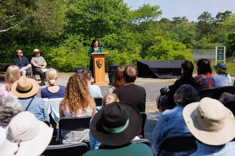 A person speaks at a podium in front of a seated audience at an outdoor installation unveiling.