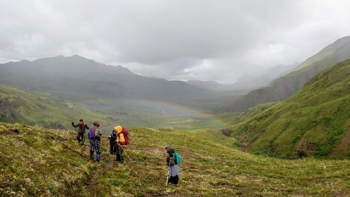five people on a mountainside looking down on a rainbow