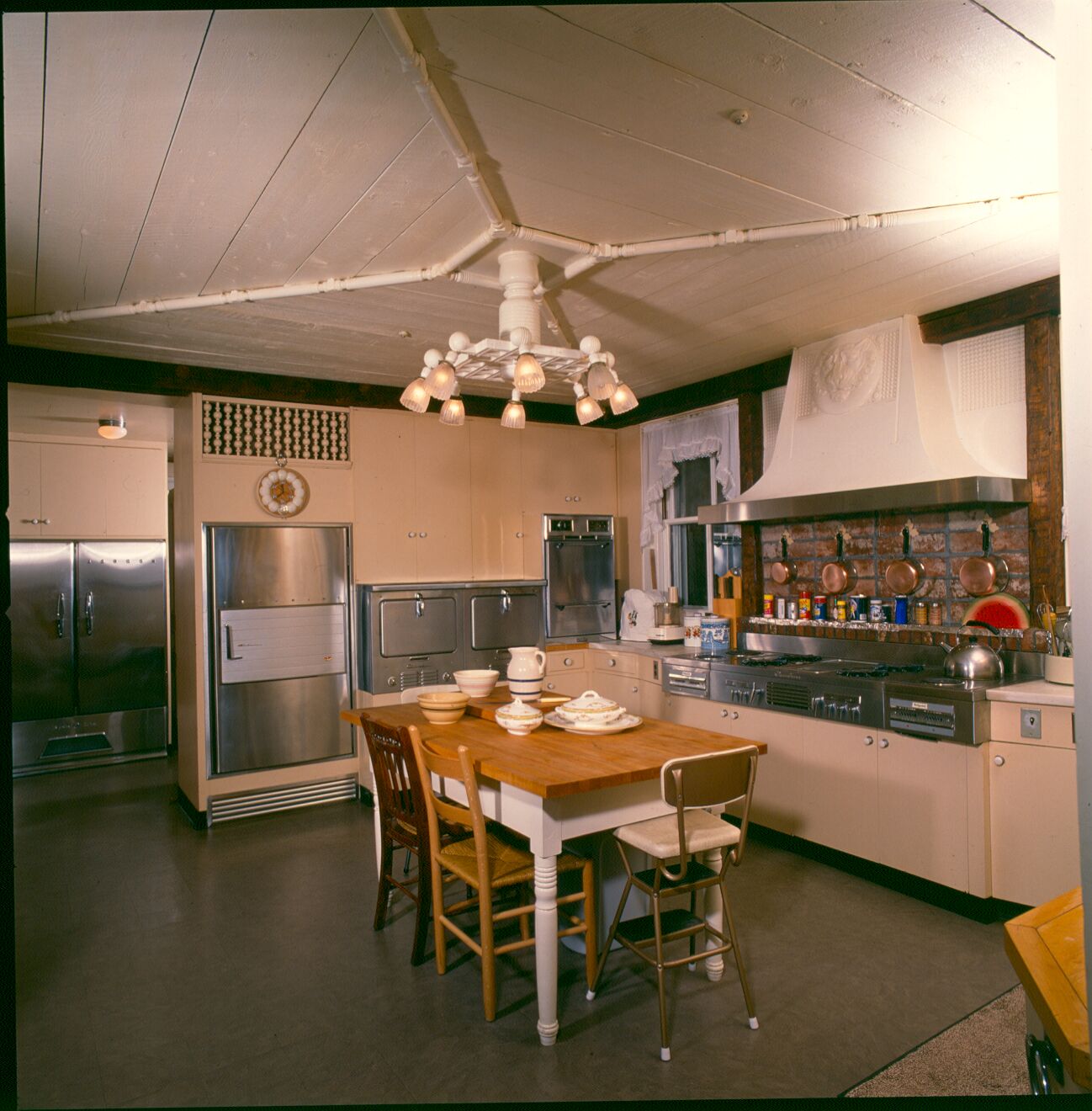 Staged photo of simple, large, 1950s-60s kitchen area with small table and chairs, white cabinets, stovetop with brick backsplash, and stainless steel appliances. 