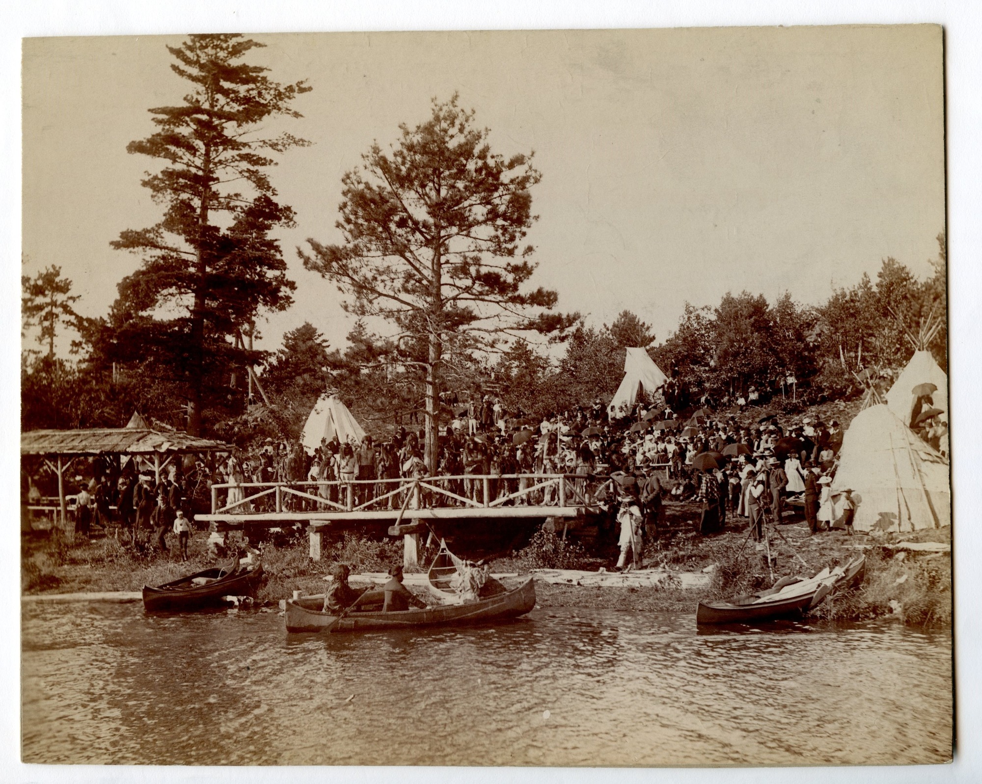 People gathered to watch production of the "Hiawatha Pageant" near shore of Lake Huron. People in boats in the foreground, people standing on a wooden bridge in middle ground, and trees and tipis in background. 
