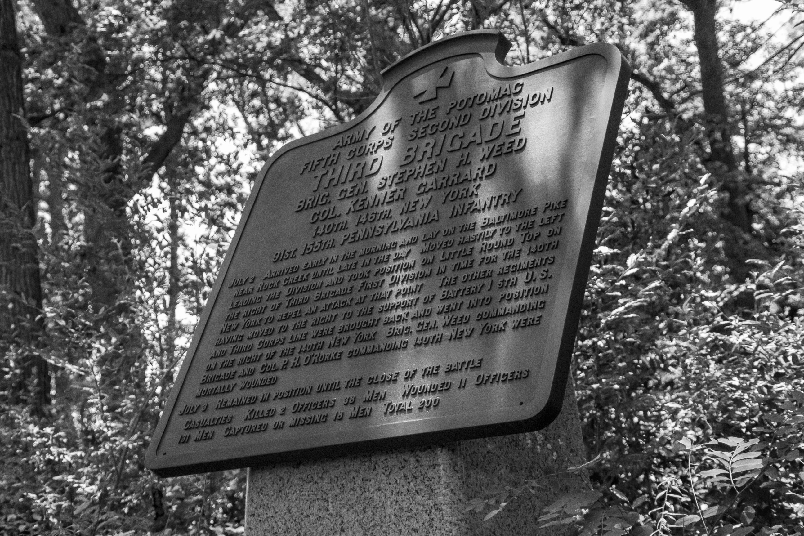 Black and white modern-day photograph of a historical marker with a stone base. metal plaque on top, and cross at the top of text describing the actions of the Third Brigade.