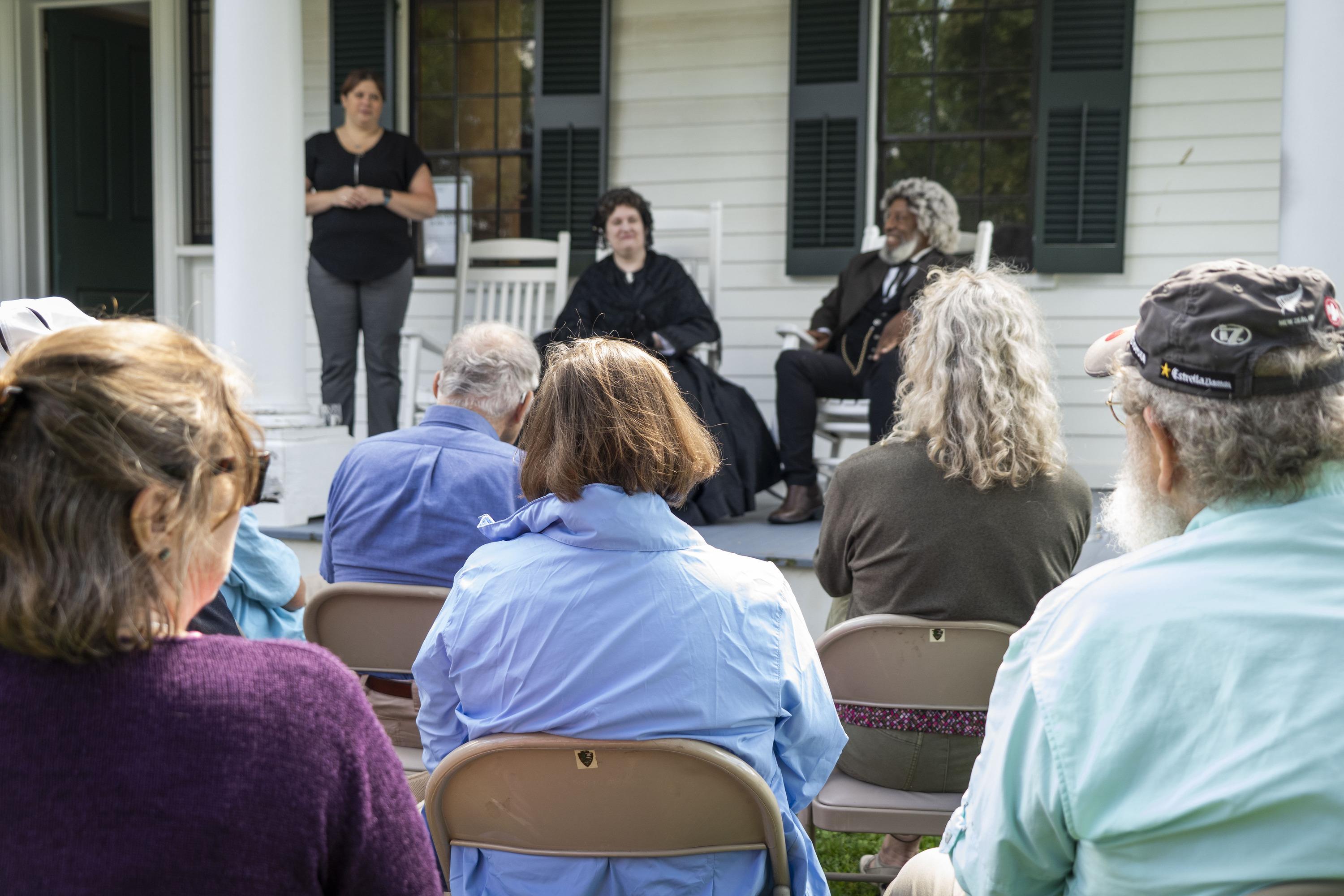 A woman and man wearing historical clothing sit in rocking chairs on the porch of a house. A group of people is gathered in chairs in front of them.