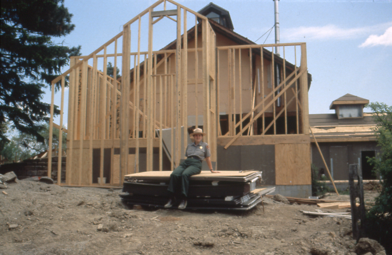 A uniformed ranger sits on a stack of plywood outside a partially complete wood-framed building attached to an existing barn.