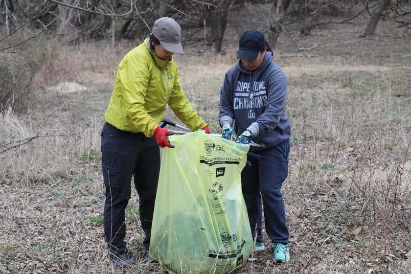 Two people wearing caps, long sleeves, and gloves are participating in a cleanup. The person on the left, wearing a chartreuse jacket and dark pants, holds open a large, yellow trash bag. The person on the right, wearing a gray sweatshirt and dark pants, is dropping trash into the bag. They are standing in a dry, grassy area with trees in the background.
