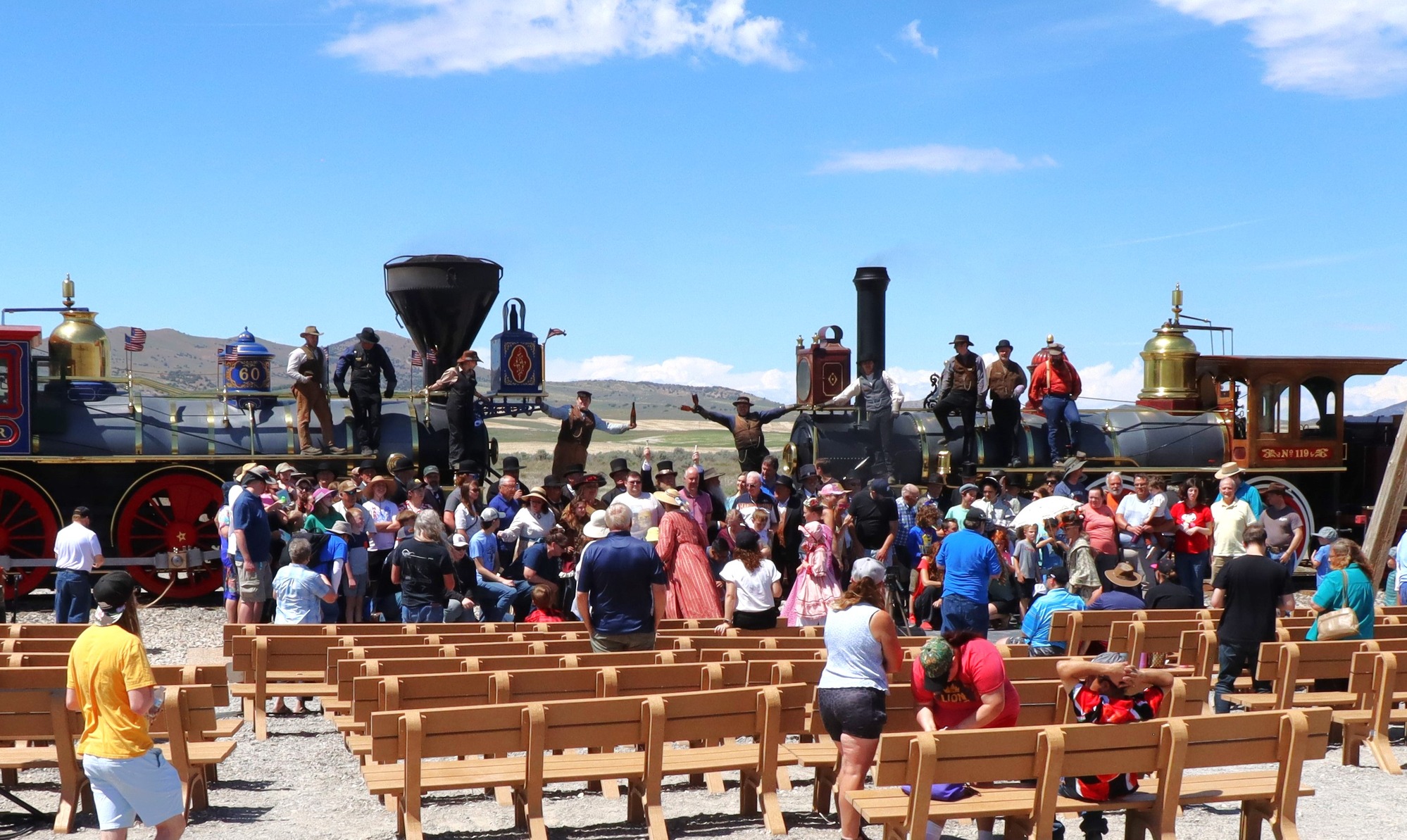 Men and women in turn-of-the-century costumes stand in front of two vibrant historic locomotives, while two engineers raise champagne bottles. Visitors in foreground taking photos.