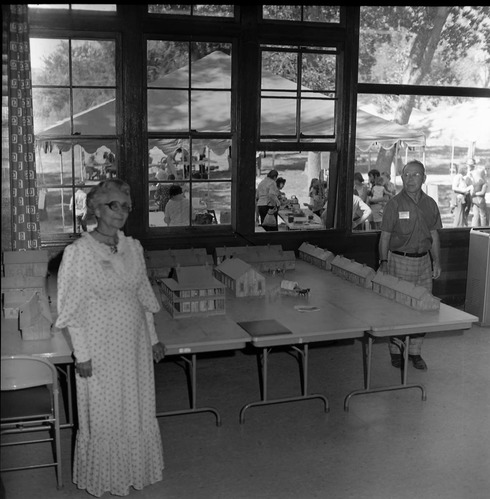 Woman and man stand near an indoors diorama of pioneer homes at the first annual Folklife Festival, Zion National Park Nature Center, September 1977. Festival activities visible outside in background.