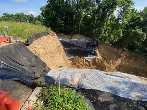 Construction site. Right side of image has a very high earthen wall with a deep excavated area continuing to the right. There are black and clear plastic tarps on either side of the excavated area. Trees and grass on the edge of the excavated area.
