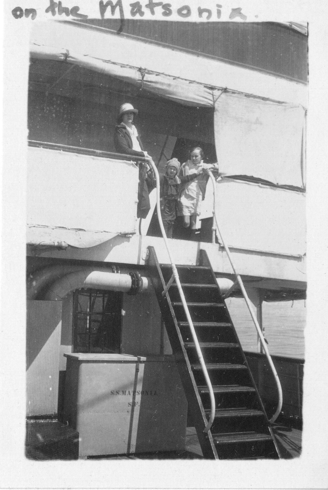 A black and white image of a woman and two children aboard a ship named, “S.S. Matsonia.” The woman and children are in the top center of the image. They are standing at the top of a set of stairs leading to a balcony. The woman is wearing a hat, a collared shirt, jacket, and pants. Her right arm is resting on top of a railing. She is looking towards the camera. The two children are to the right of the woman. They are both wearing jackets and a dress. The child closest to the woman is holding onto the top of the stair railing with their left hand. The second child is older than the one holding onto the stair railing. She is also holding onto the stair railing but is looking down towards the younger child. There are sunscreens attached to the roof of the balcony.  On the first floor of the ship there is a storage cabinet to the left of the stairs. There is a label on the front of the cabinet that reads, “S.S. Matsonia S.F.” Along the top edge of the photo, “on the Matsonia” is written in marker.