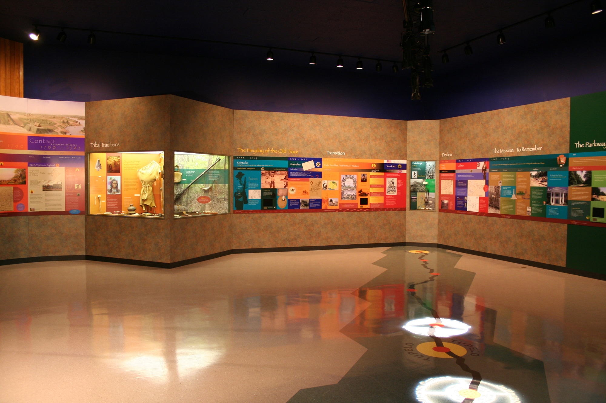 A full color photograph inside a building. The floor reflects some of the overhead lights. In the left side of the frame is a display case with some historical artifacts. Information displays line the walls, but the text is not legible in this photo. On the floor coming from the bottom right of the frame are tiles painted to mimic a map of the Natchez Trace Parkway.