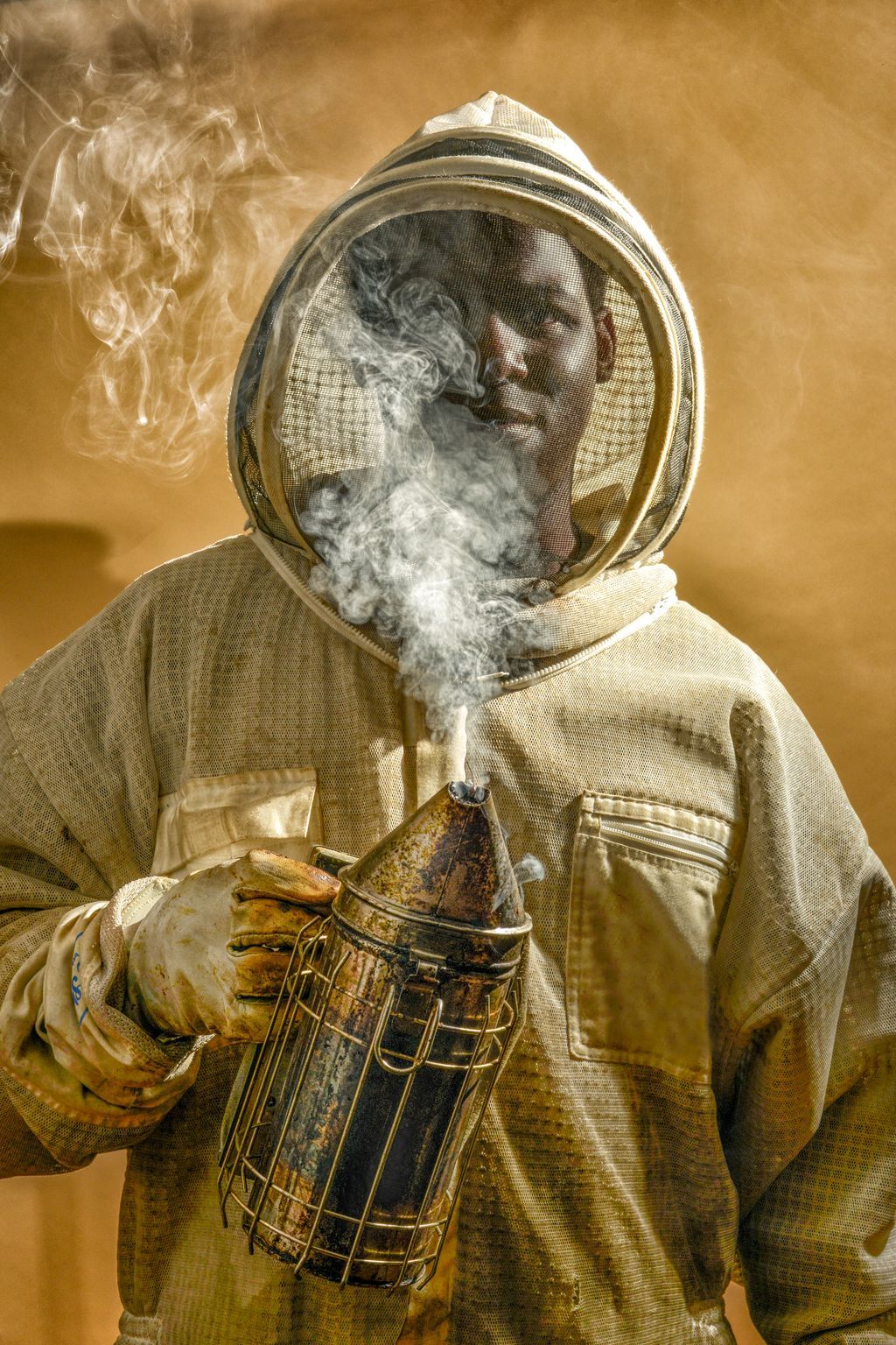 A man stands in a bee-keeping suit, holding a smoker in one hand that wafts smoke up past his face.