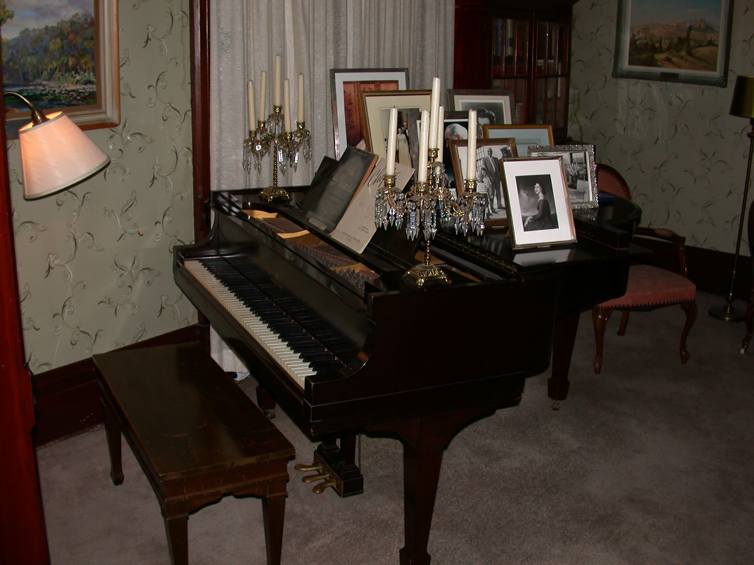 The music room/ parlor. A baby grand piano is adorned with eight photographs on top. A desk in the background has several books. Candelabras adorn the piano.
