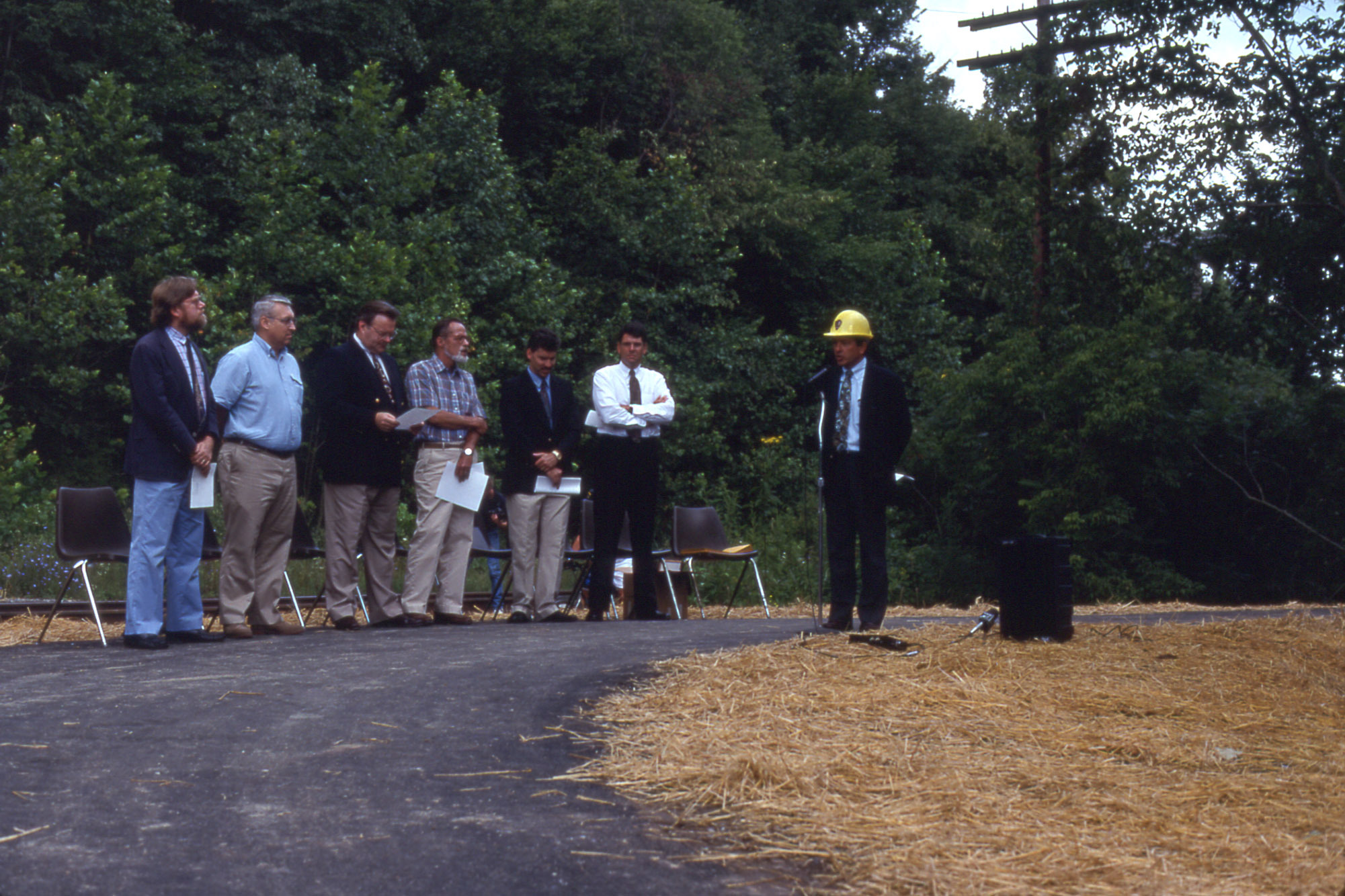 A man in a hard hat stands on a grey path speaking into a microphone as six men stand holding papers in front of a line of chairs. The ground is covered in straw.