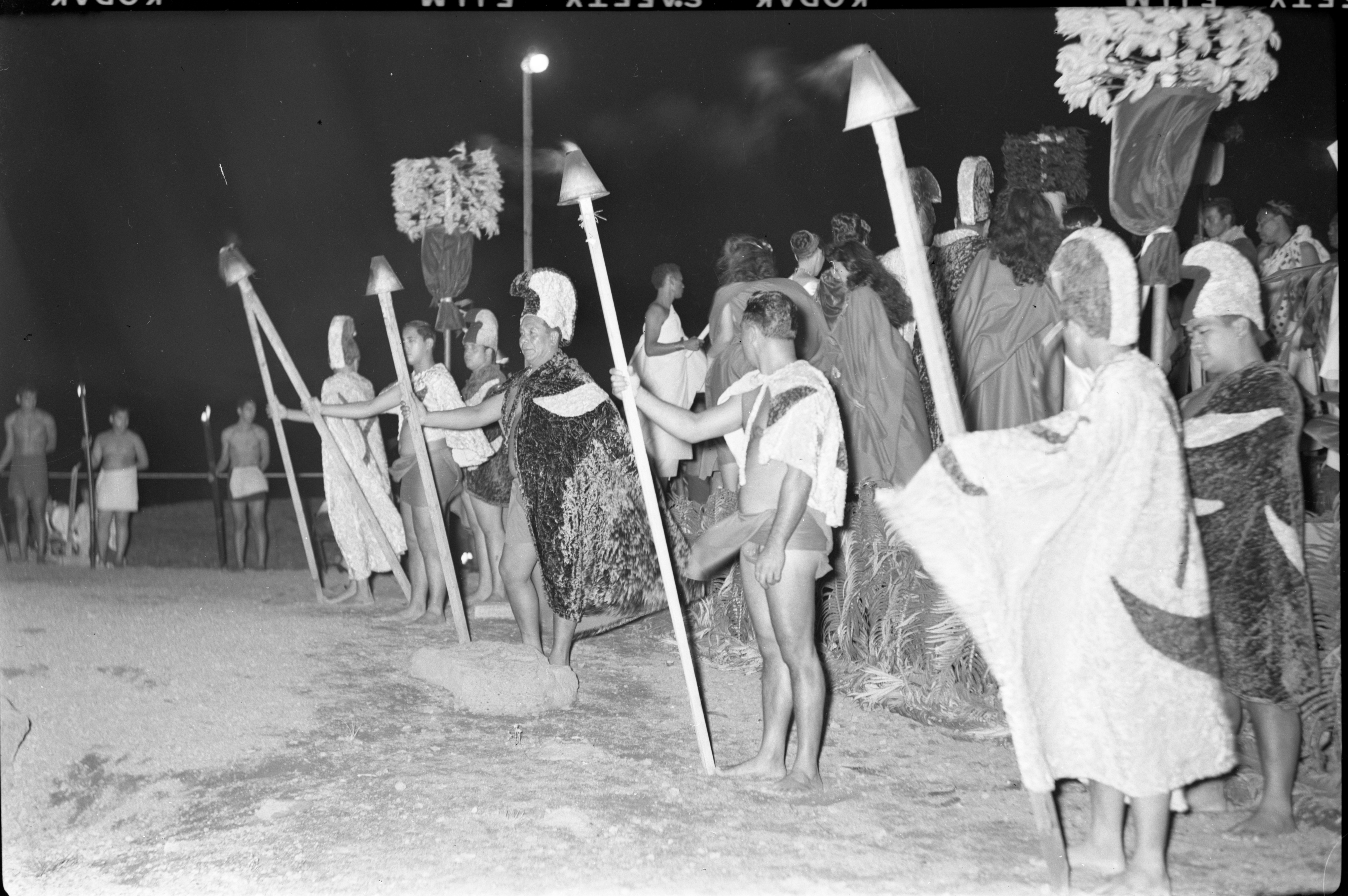 A black and white image of a line of men holding bamboo torches during a pageant at Halemaʻumaʻu crater. There are five men equally spaced standing in a row facing the left side of the image. Each of the men are holding a lit bamboo torch in their right hand. Three of the men, one being closest to the right side of the image, then the third and sixth from the right, are wearing a mahiole, ʻahuʻula, and malo. The second and fourth man from the right side of the image are wearing ʻahu and malo. Behind the first and sixth man in the row a man is holding a kāhili. They are dressed in a mahiole, ʻahuʻula, and malo. There is a fern leaf lined stage behind the men standing on the ground. There are many people on the stage with their backs facing the camera. In the background, a streetlight is visible as well as three other men in malos holding lit bamboo torches.