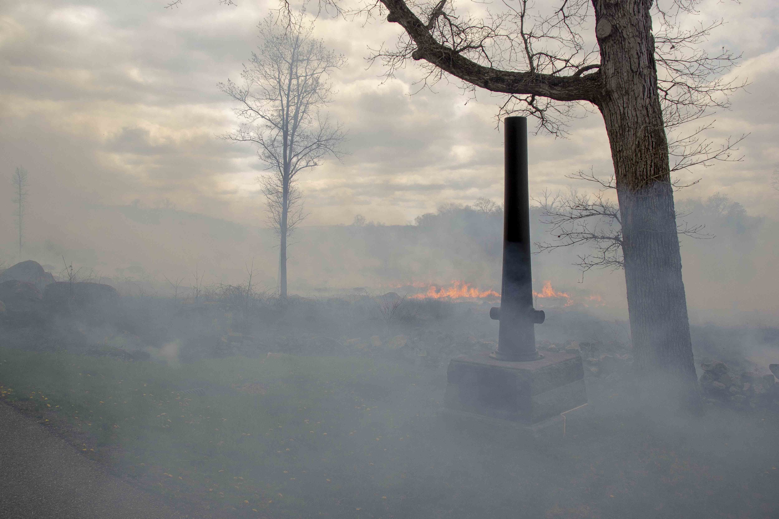 A monument with an upturned cannon embedded in a block of stone stands next to tree in front of a low stone wall. Fire and smoke rises from beyond the stone wall.