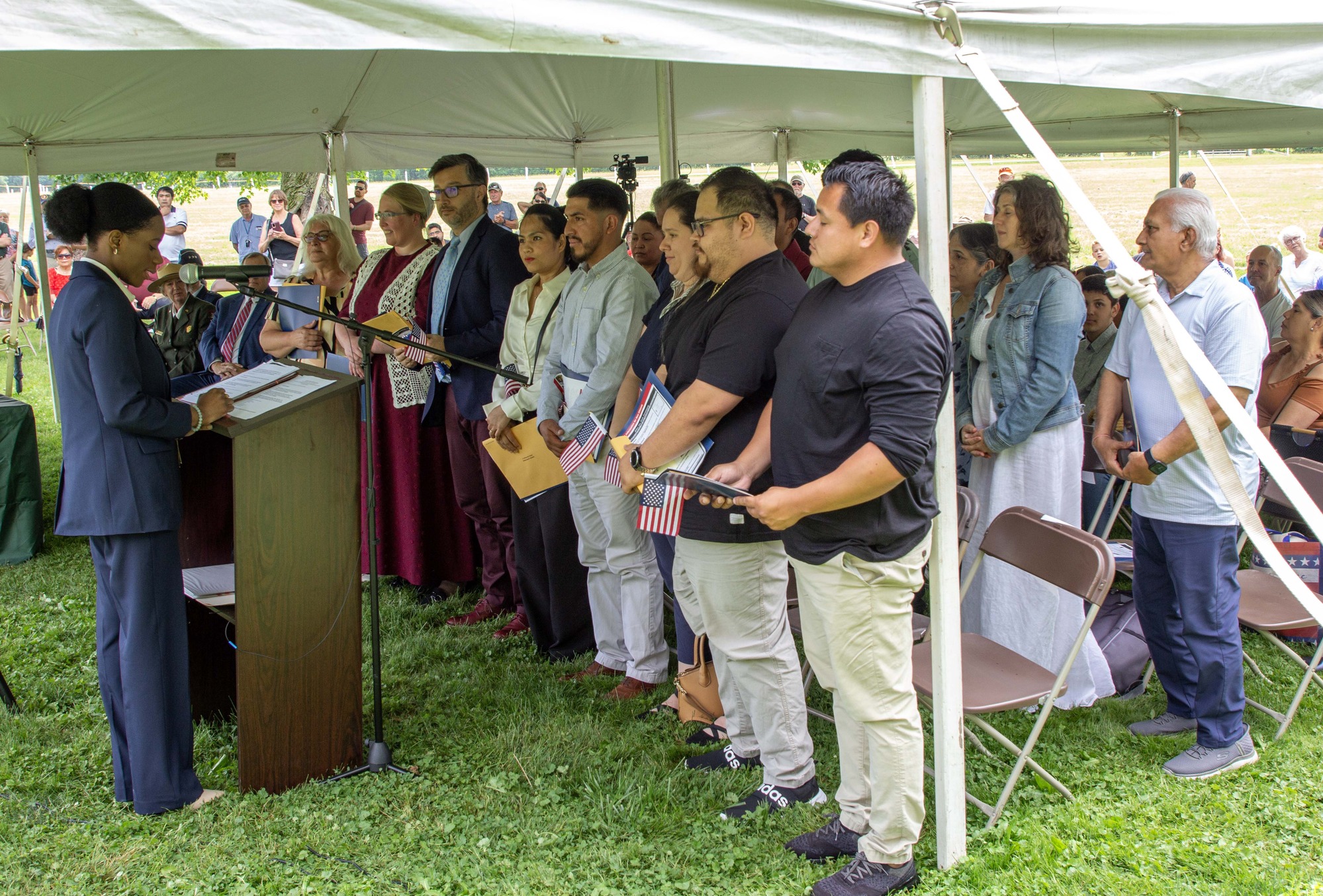A woman in a blue suit stands at a podium, with several rows of people standing in front of her. 