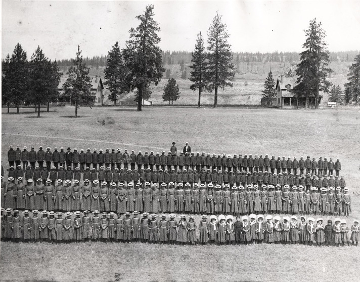 Black and white photograph of four lines of school children in matching clothing standing in a field for a photo