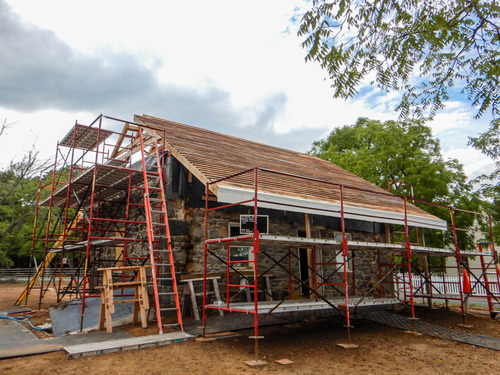 Red scaffolds and ladders are wrapped around the front and side of the house to work on the walls and roof. Light tan colored cedar shingles are on the roof which contrasts with the bright white and blue sky. 
