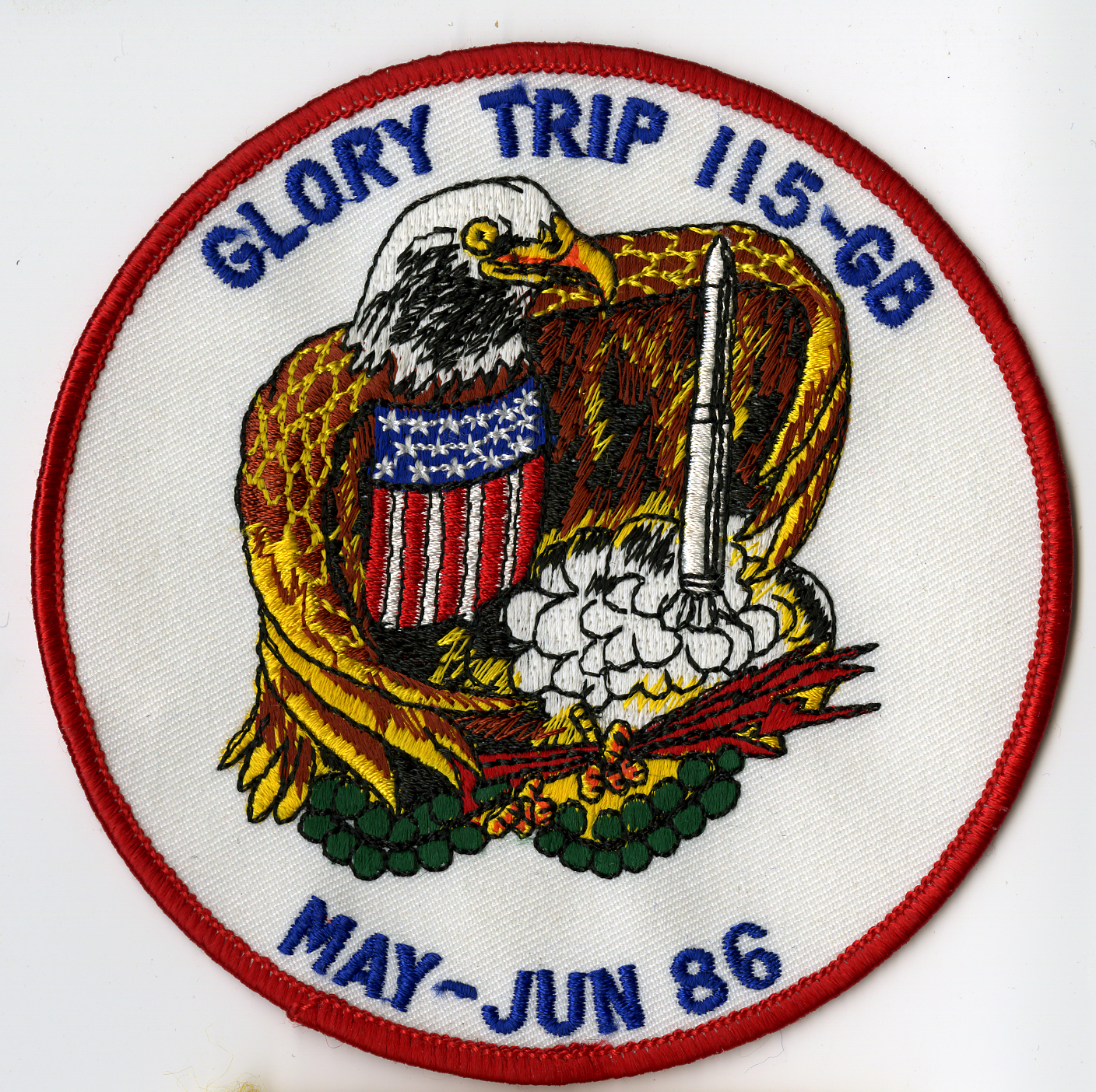 White circle with red merrowed edge. Center is pictured a bald eagle with red, white, and blue shield on chest holding red lightning bolts and green olive branches. Cradling white missile with white and yellow smoke plume coming from bottom of missile. Around edge is GLORY TRIP 115-GB / MAY-JUN 86 in blue.

