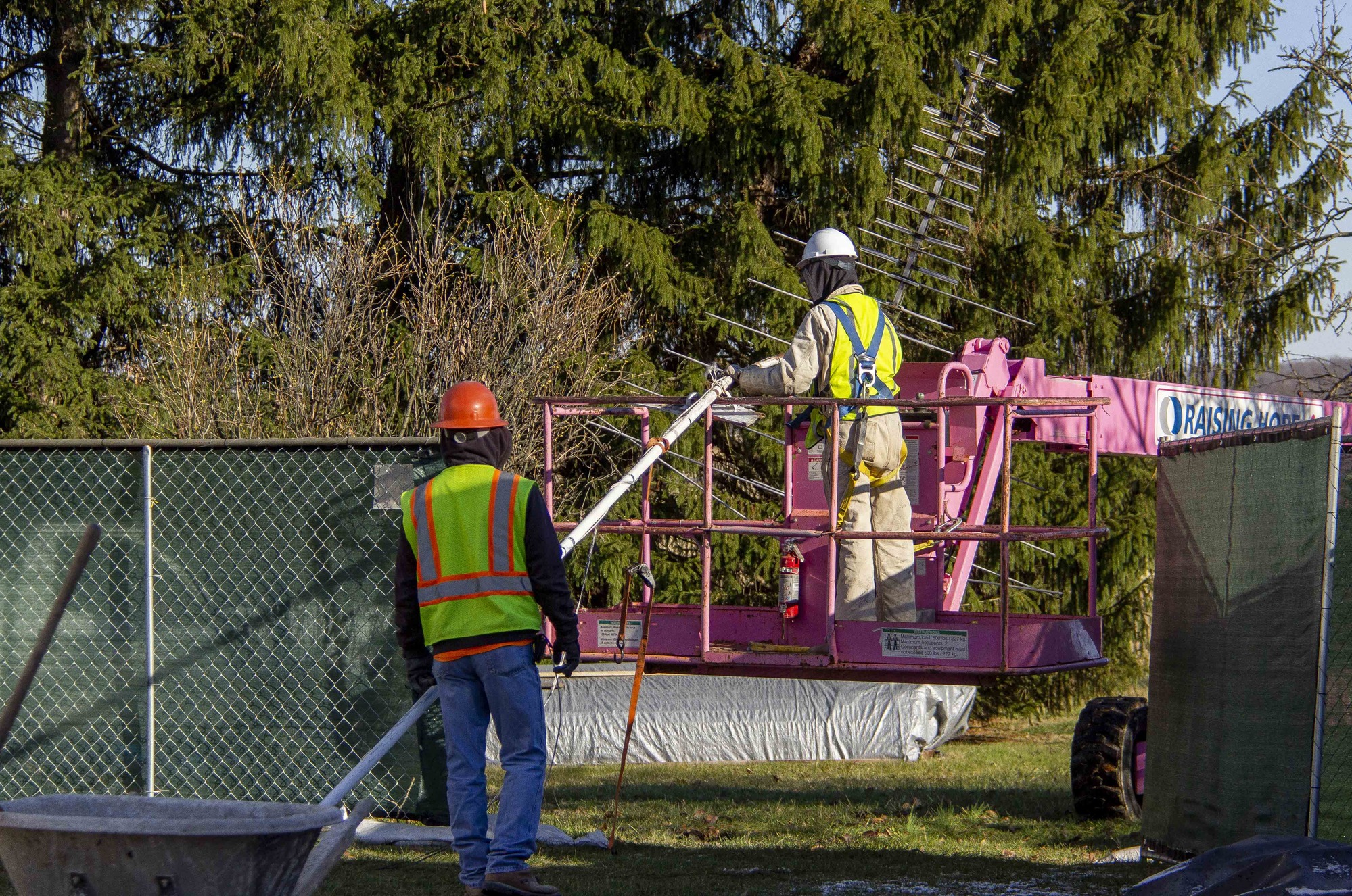 Two construction workers continue the process of removing the old TV antenna from the side of the house. The worker navigating the antenna down in the pink lift is almost at ground level with the second worker. 