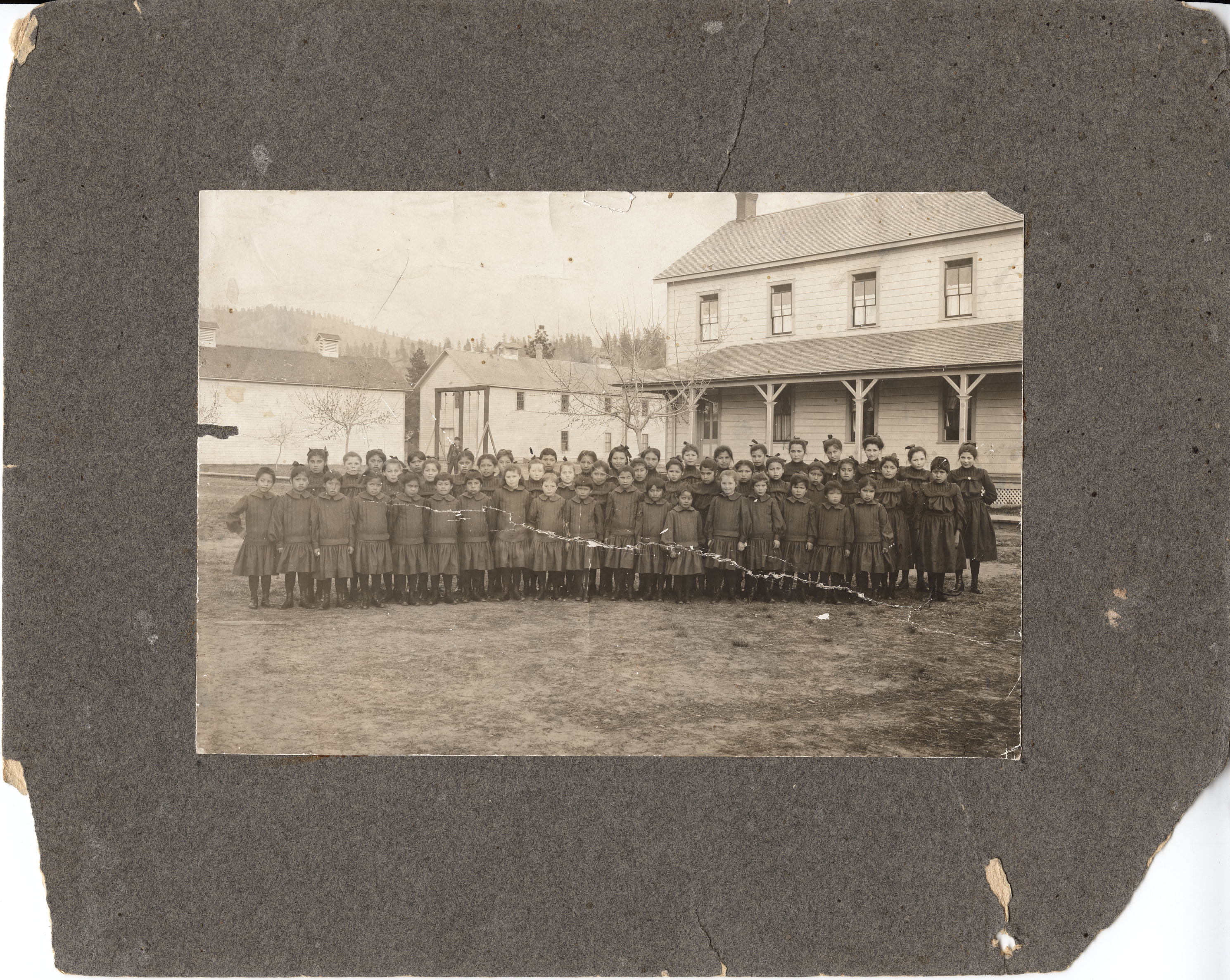 Black and white photograph with wide border of several rows of identically dressed American Indian students standings in front of buildings