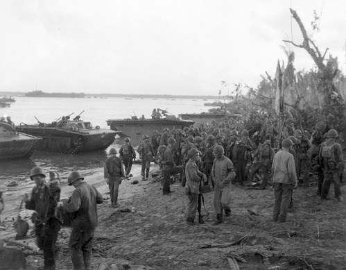 Soldiers in military gear gather on a muddy beach, with landing crafts in the background and a cloudy sky above.