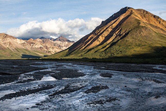 a large mountain covered in green with more peaks in the background and a river in the foreground
