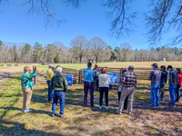 A park ranger talks to a group of people overlooking a field with a wooden fence and a small log cabin in the distance
