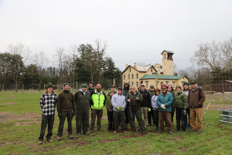 A group of 19 people in hats, coats, and work clothes pose in the orchard, with the two and a half story mansion with tower in the background. 