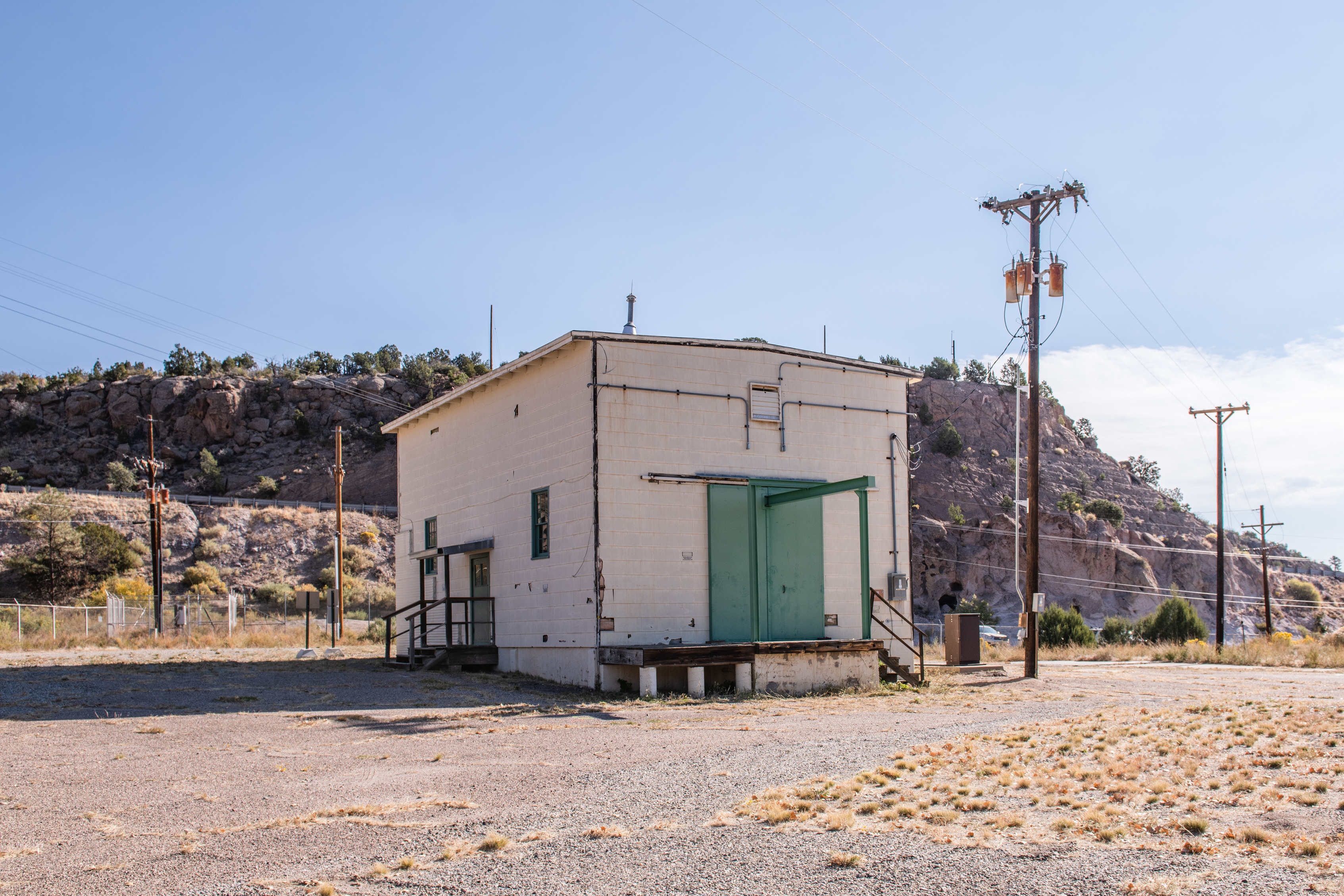 A white square building with teal steel entry doors, the Slotin Building, stands connected to power lines and poles in a dry, flat field with a rocky, high desert ridge behind.