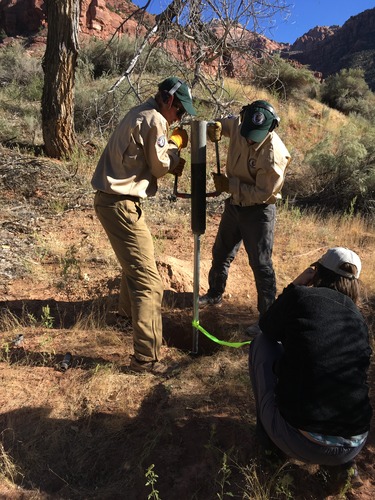 Two young men in green ball caps and khaki uniforms hold either side of a well installation device. 