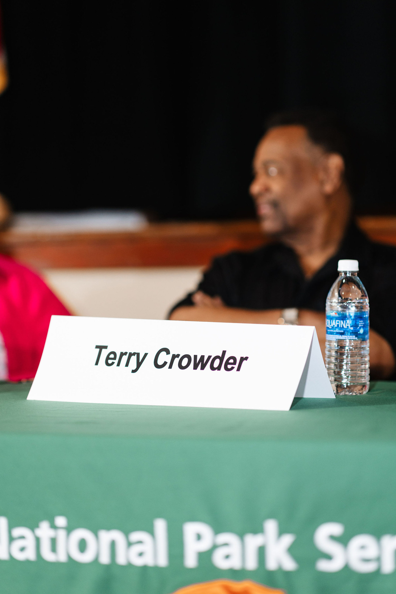 An out of focus African American man in a black shirt behind an in focus table tent reading terry crowder crosses his arms as he looks at the panelist next to him.
