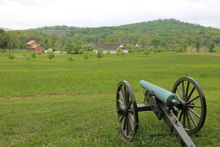 A teal-colored tarnished bronze cannon looks out across a green field with small trees in the distance. A brick home and white barn are in the distance and there are two hills with forested areas in the upper portion of the photo. 