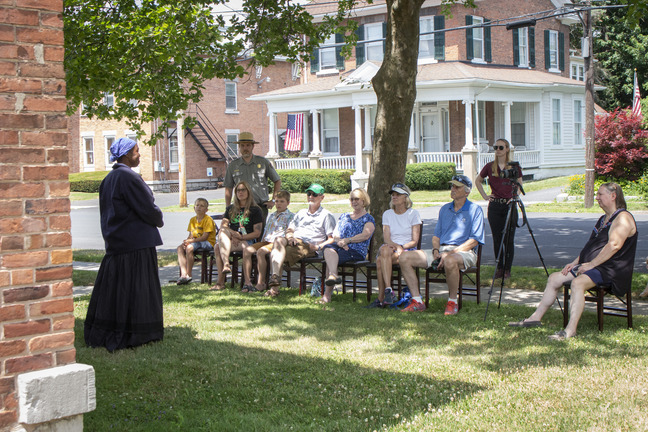 A woman stands in a long black dress in between a house and a small group of seated people.