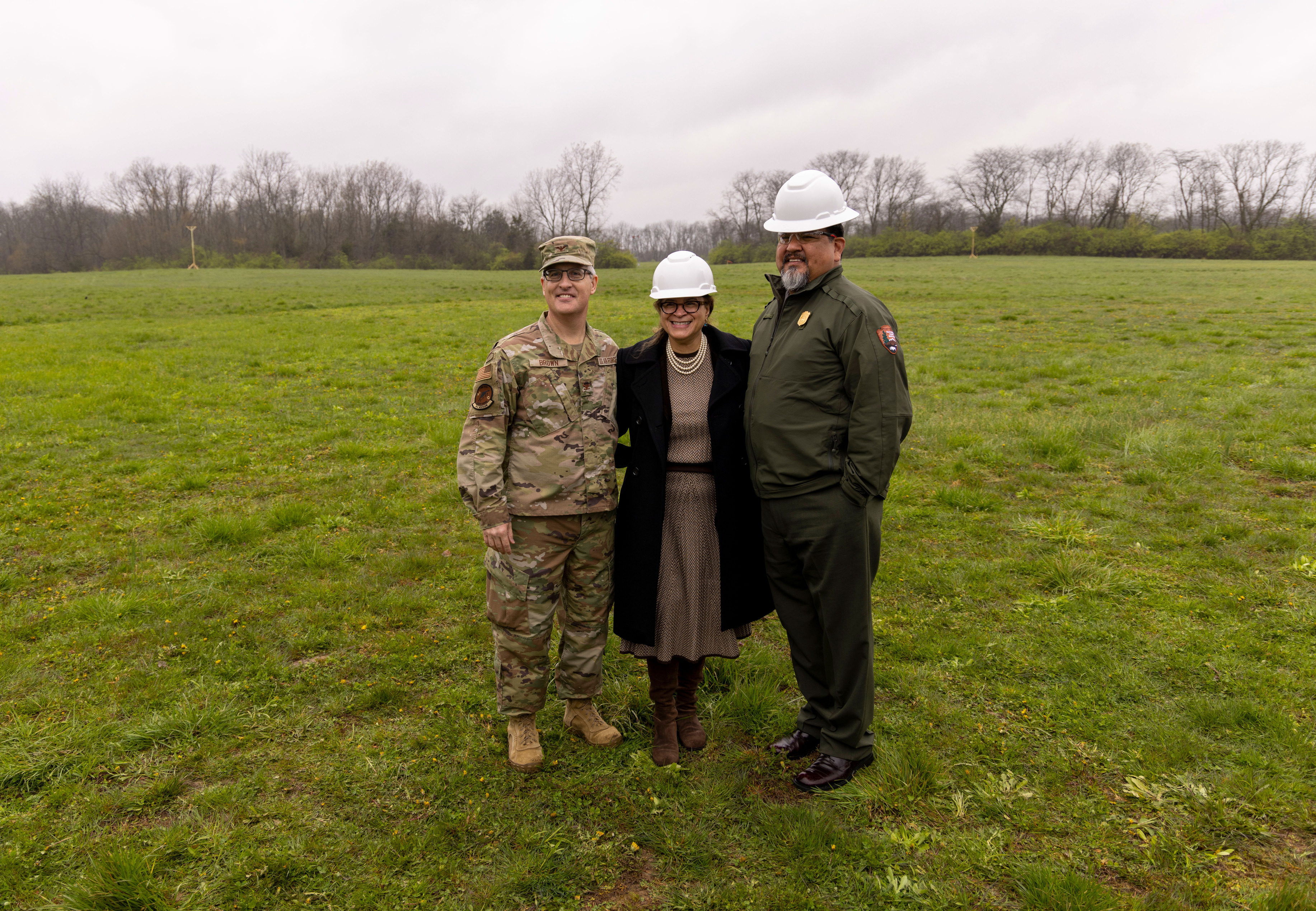 A man in a white hard hat and green uniform stands next to a lady in a brown coat and white hard hat and a man in a camouflage uniform