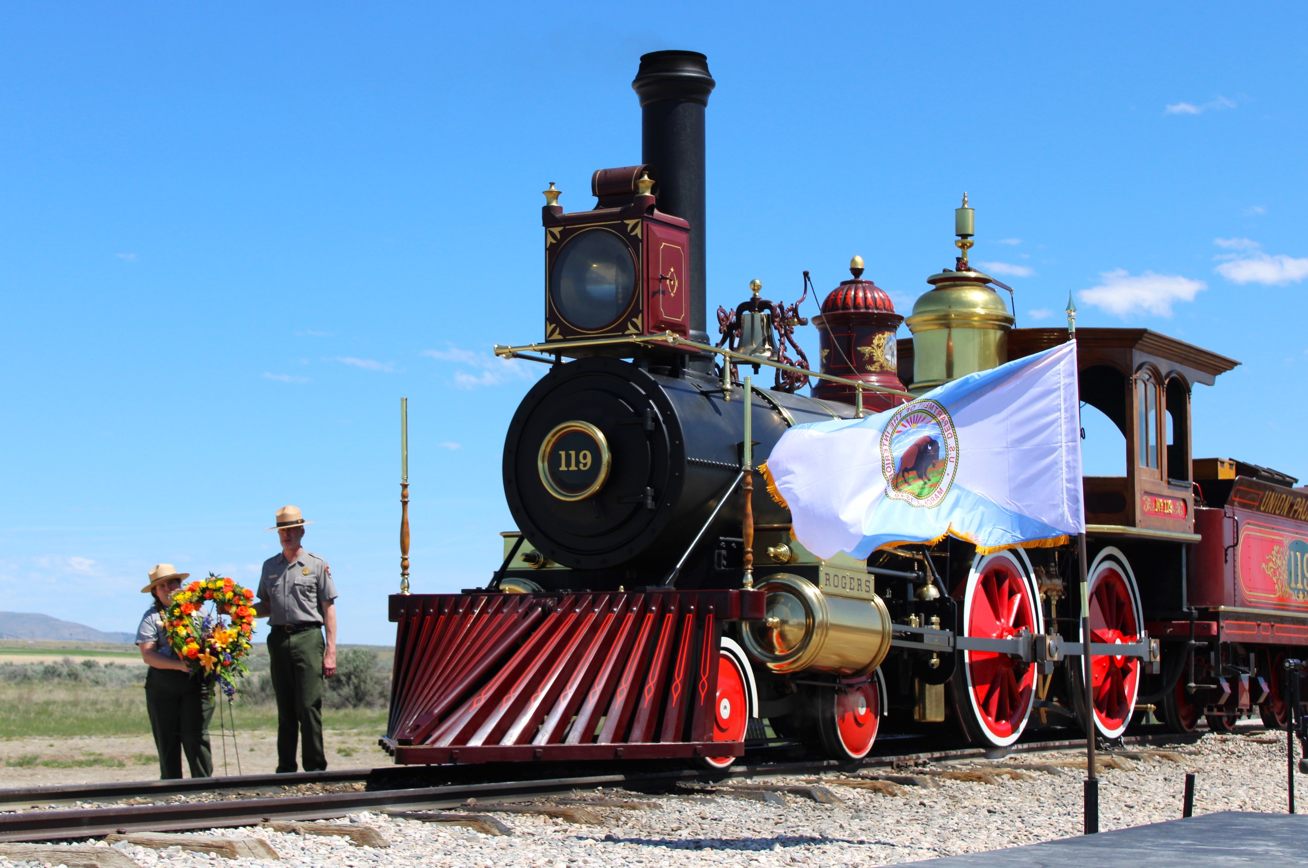 Two rangers stand behind railroad tracks along side the 119 Locomotive with a standing Wreath of Flowers.
