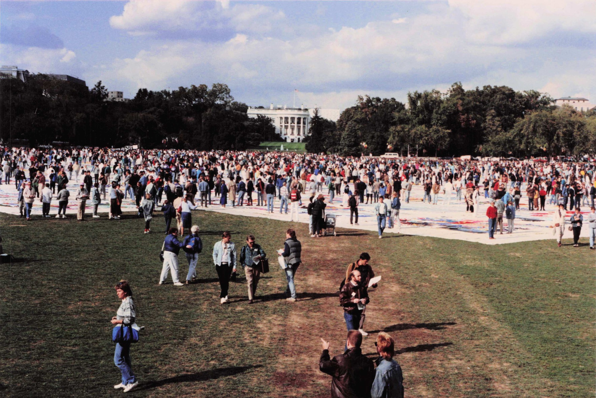 A collection of people stands on multi-colored cloth laid on the grass. They are looking down at the cloth. Trees and the south lawn of the White House are visible in the background