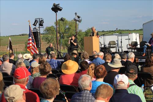 A crowd of people sit outside watching a speaker on stage. A sign language interpreter is sitting to the left.