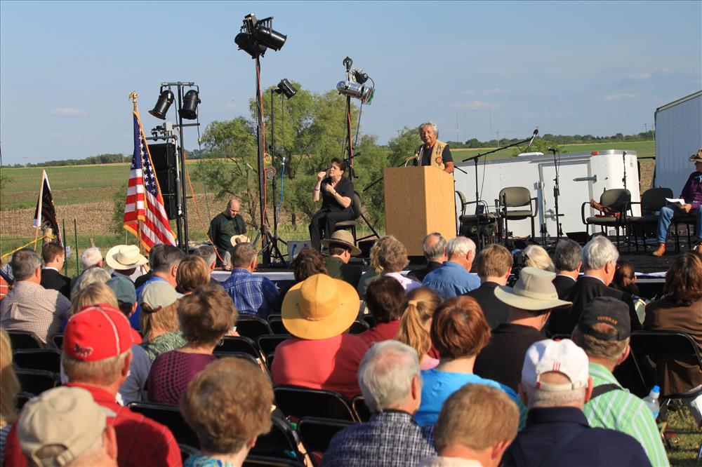 A crowd of people sit outside watching a speaker on stage. A sign language interpreter is sitting to the left.