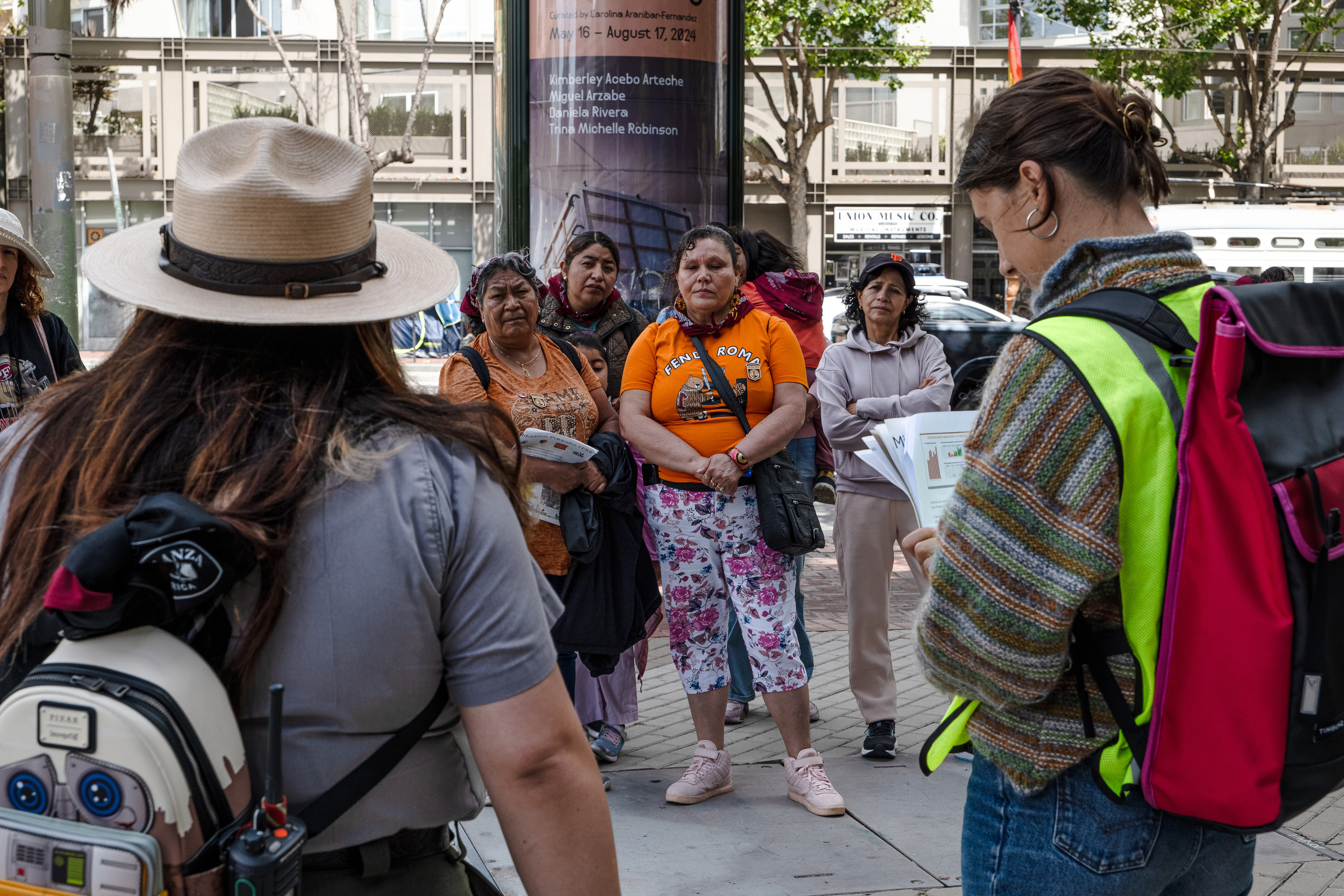 A group of women and children gather together on a city sidewalk to listen to a speaker wearing a high vis vest and an NPS Park Ranger