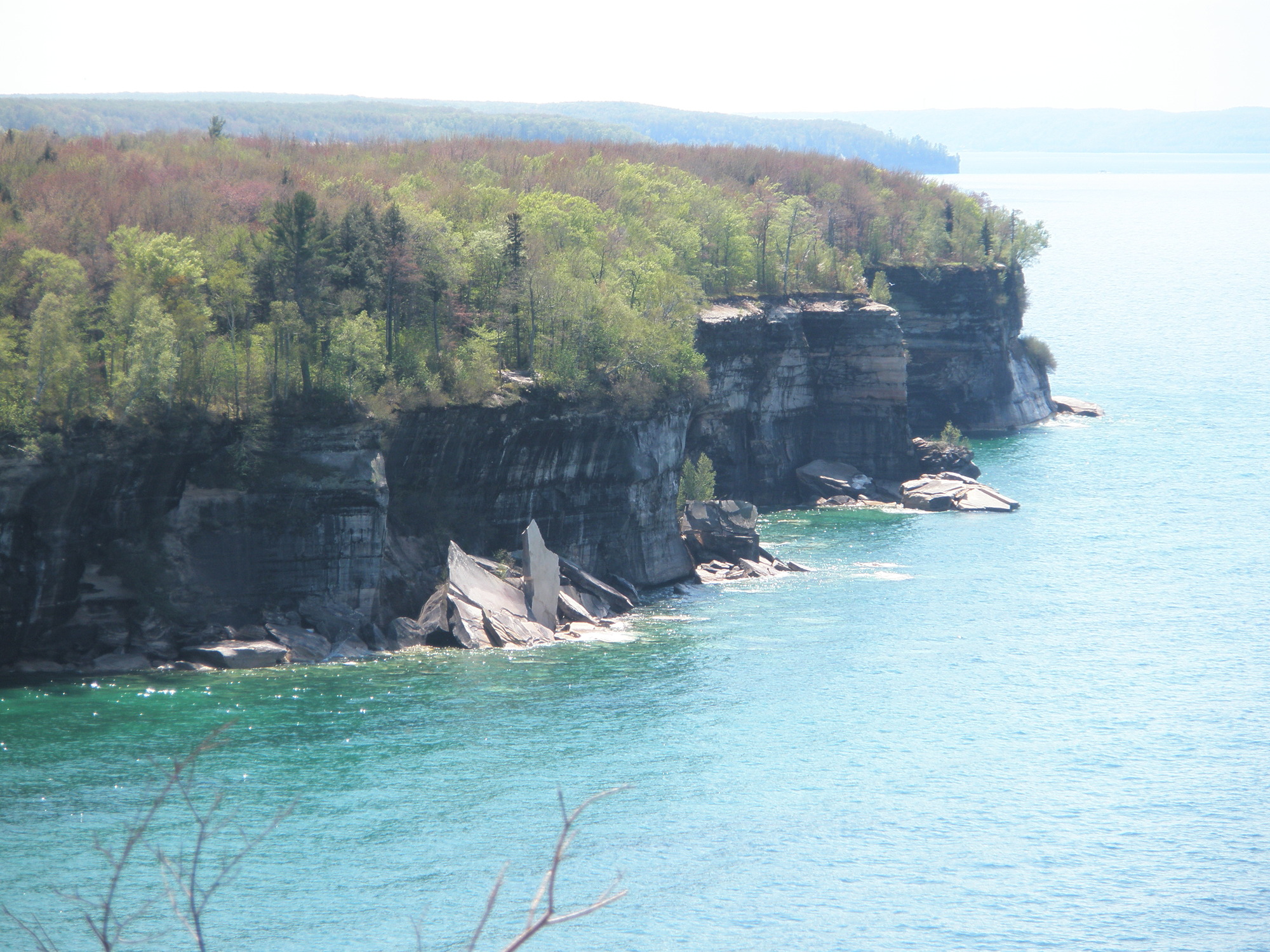 Black-streaked cliffs rise out of Lake Superior. Broken cliff sections sit at the base. Trees are growing on top.
