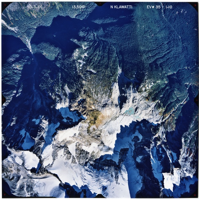 Color aerial photograph of glacier and snowfields on mountain peak, with forested valleys below peak.