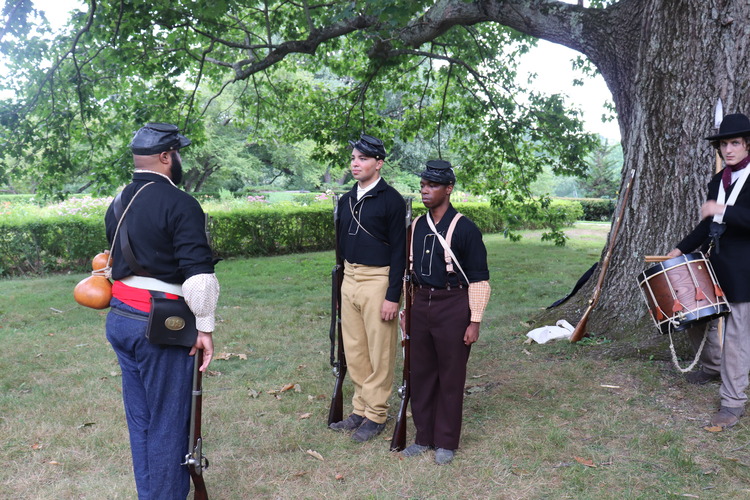 3 black militia men drilling in front of a large tree while a white drummer looks on. 