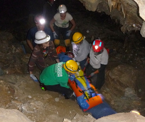 Cavers in helmets gathered around an individual wrapped in a splint within a cave cavity.