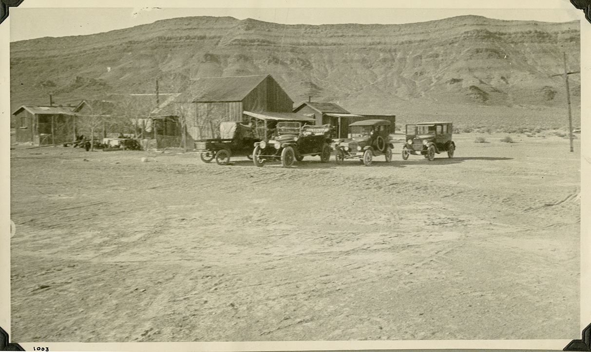 This is an historic black and white photograph from the Scotty's Castle Historic Photograph Collection, Death Valley National Park of four automobiles parked near wooden structures, possibly in Bonnie Clare, Nevada. Circa 1919, the year Johnson took or had someone take many photographs prior to construction.