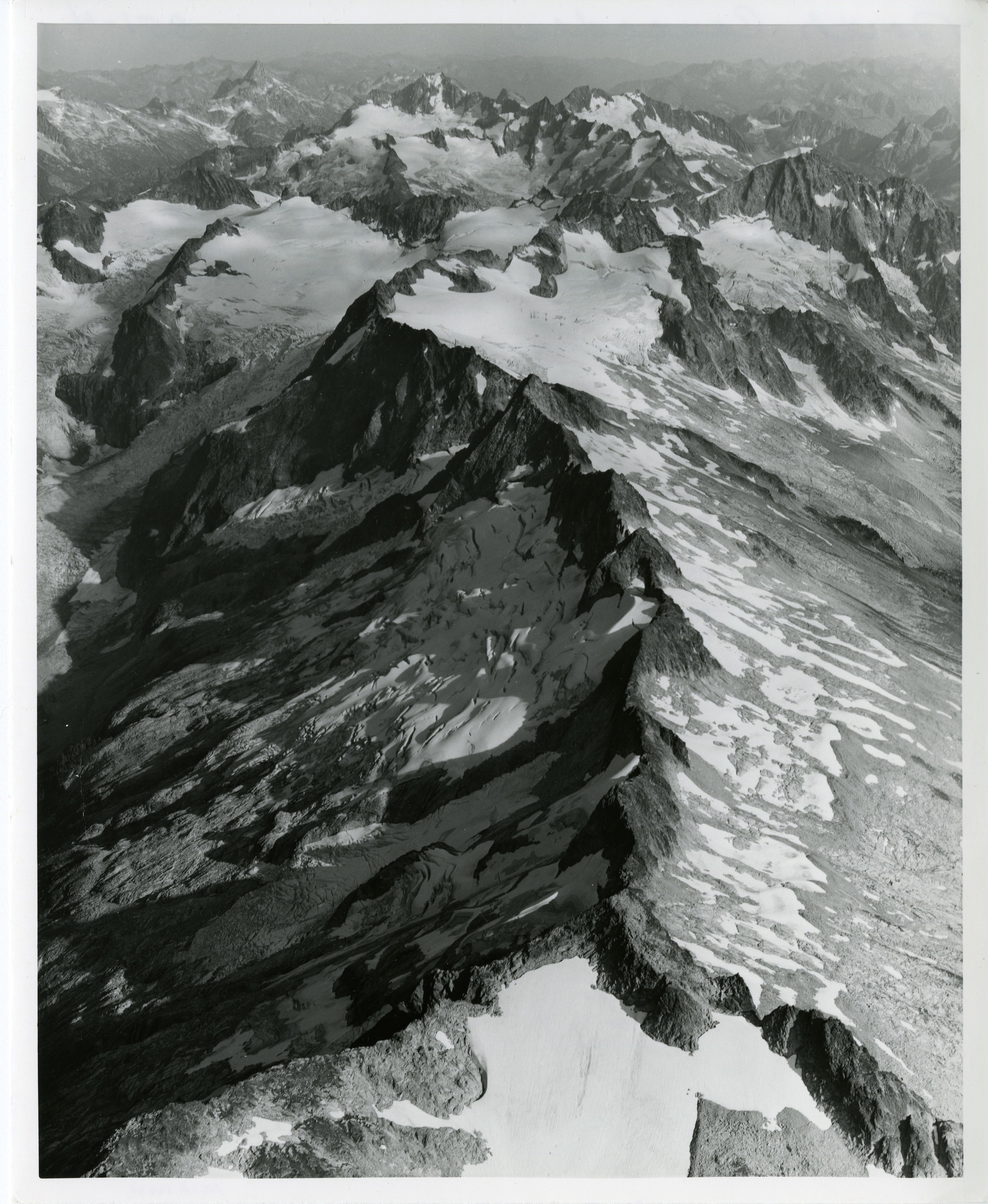 Aerial view of the top of a mountain peak, covered in snow and glaciers. More peaks stretch to the horizon.