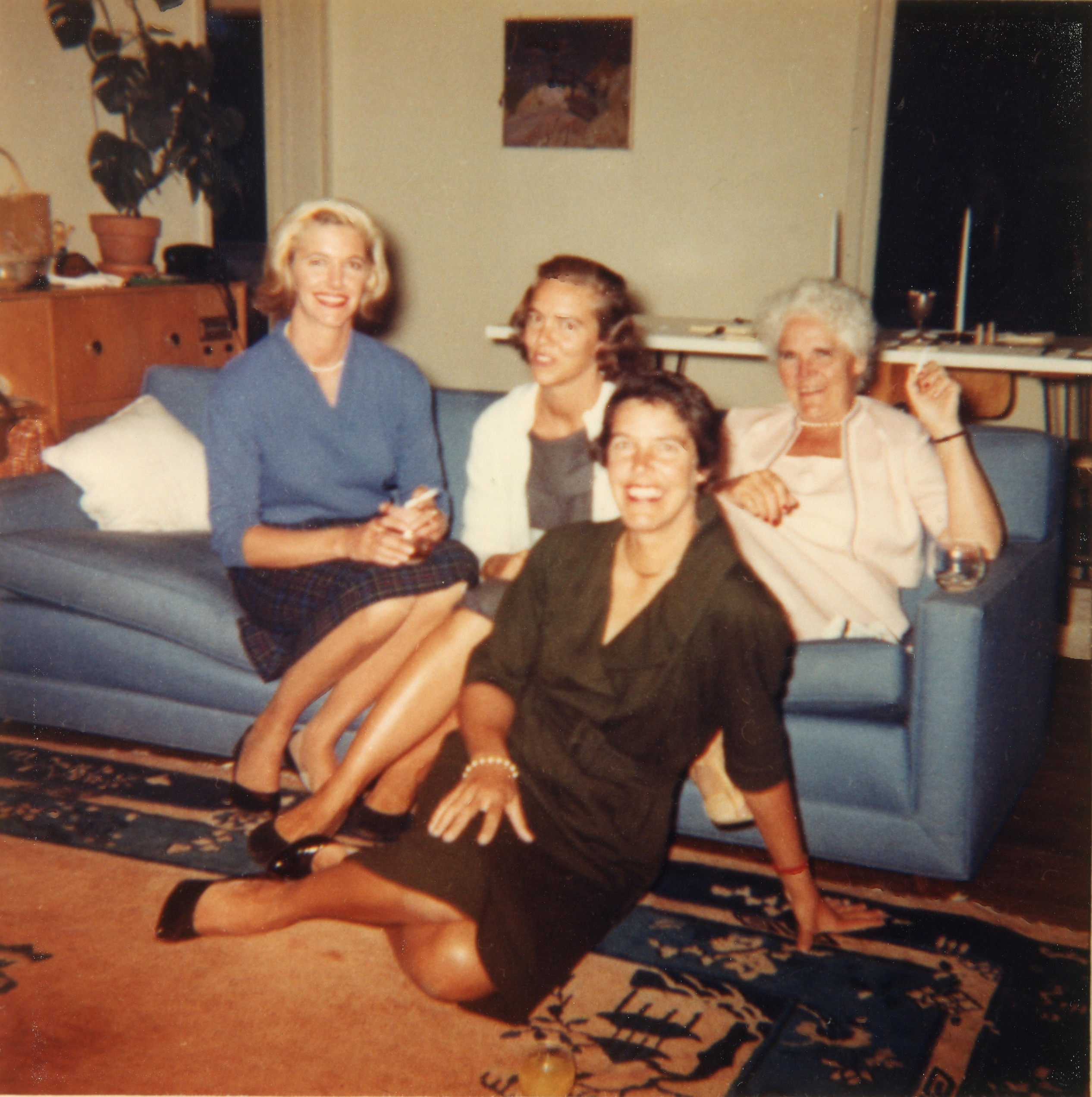 A color photo of four women. Three women are sitting on a blue couch with one woman sitting in front of the couch. 
