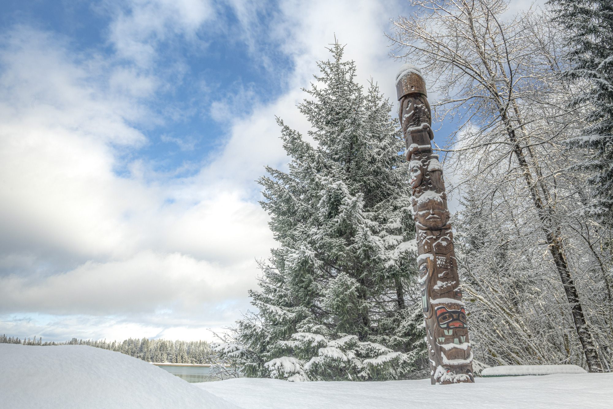 The healing totem pole in Bartlett Cove, covered in light snow on a partly sunny day, blue sky peeking out between fluffy clouds. Surrounding trees and the ground are covered with snow.